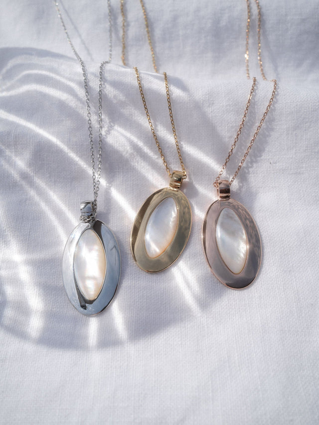 Island Elegance Kette von PURELEI mit ovalem Perlmutt-Anhänger in Silber, Gold und Roségold auf weißem Stoff.
