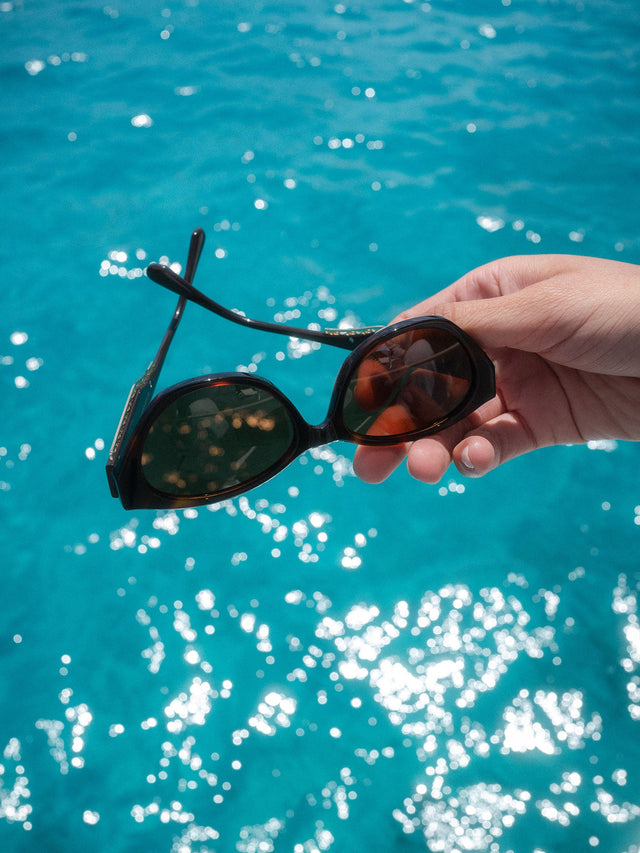 Lagoon Sonnenbrille von PURELEI, dunkelbraun im ovalen Schildpatt-Design mit goldenen Details vor blauem Wasser.