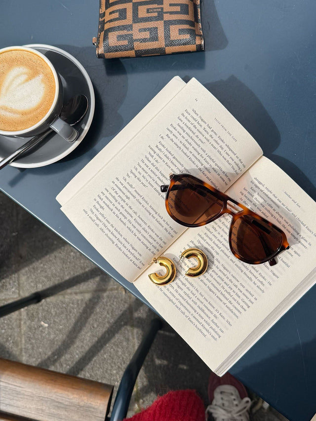 Palm Beach Sonnenbrille von PURELEI in braunem Schildpatt mit goldenen Bügeln auf offenem Buch, warme Sommerstimmung.